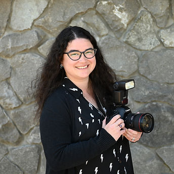 Smiling woman holding a camera, wearing glasses, in front of a stone wall. theshutterbug staff kyla